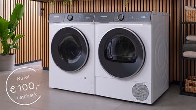 Twee witte Siemens wasmachines staan naast elkaar in een wasruimte met houten lattenwerk aan de wand. Links staat een grote groene plant in een grijze pot, rechts zijn rieten manden op planken te zien. Op de afbeelding is een promotieaanbieding zichtbaar met "Nu tot €100,- cashback" in een cirkel linksonder