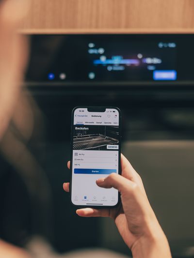 Eine Person steht in der Küche vor einem Backofen und bedient die Home Connect App auf einem Snmartphone.