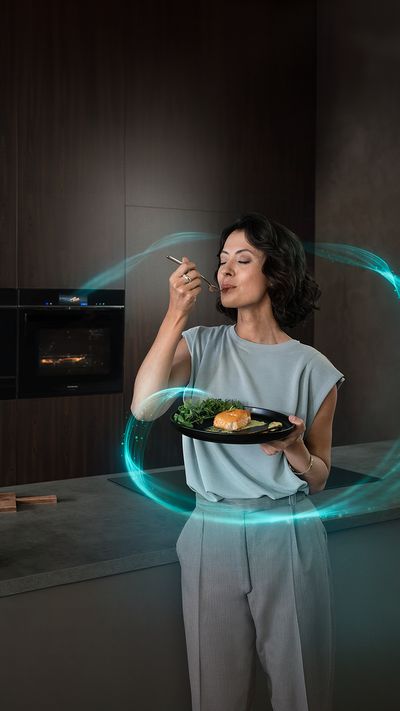 Eine Frau steht vor einem eingebauten iQ700 Backofen. Sie hält einen Teller mit einem Lachsgericht und eine Gabel in der Hand.