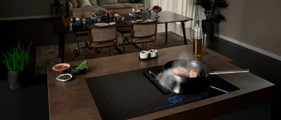 Cucina moderna con piano cottura a induzione, padella per cucinare e tavolo da pranzo con sedie e stoviglie sullo sfondo. 
