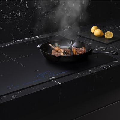 Una fetta di carne che sfrigola in padella su un piano a induzione elegante di colore nero, avvolta da una nuvola di vapore, con varie fette di limone su un tagliere.