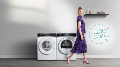 Eine Frau in einem lila Kleid geht neben einer modernen Waschmaschine und einem Trockner vorbei, an deren Wand eine Cashback-Aktion abgebildet ist.