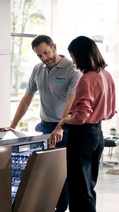 Ein Servicetechniker erkl&auml;rt einer Kundin einen ge&ouml;ffneten Siemens Einbau-Geschirrsp&uuml;ler in einer modernen K&uuml;che.