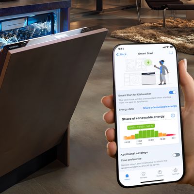 En person håller en smartphone som visar diskmaskinens inställningar med energidata, bredvid en öppen diskmaskin i ett modernt kök.