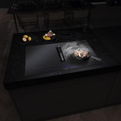 Un piano di lavoro nero dall'eleganza contemporanea, con una fetta di carne saporita che sfrigola e cuoce in padella su un piano a induzione, pronta per essere impiattata con cura tra varie fette di limone.