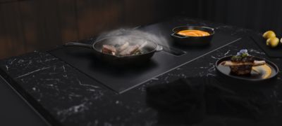 Een pan met gebakken biefstuk en kruiden, met daarbij een schaal met romige saus op een donker marmeren aanrecht. 