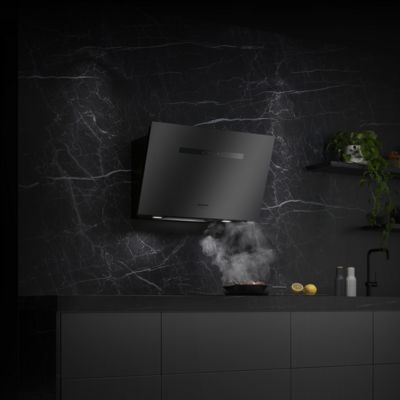 Una cappa elegante di colore nero su una parete scura in marmo, con un filo di fumo che sale da una padella sul piano di lavoro e varie fette di limone sul ripiano.