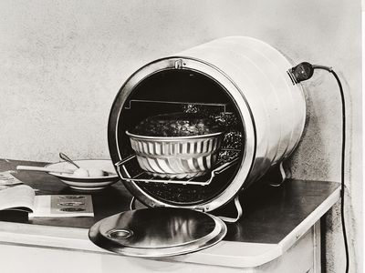 Dobov&aacute; elektrick&aacute; trouba s obl&yacute;m kovov&yacute;m proveden&iacute;m a vložen&yacute;m pek&aacute;čem stoj&iacute; na stole vedle kuchařky a tal&iacute;řů.