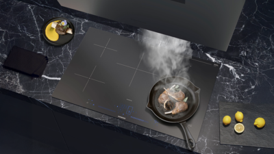 Matte K&uuml;cheninsel in dunkler Optik mit Siemens Induktionskochfeld und integriertem Dunstabzug; Dampf steigt aus einer Pfanne auf, warmes Licht im Hintergrund.