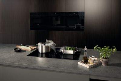 A black Siemens induction cooktop with an integrated downdraft extractor actively pulling steam from a pot and a pan of vegetables, set in a modern kitchen with dark cabinetry.