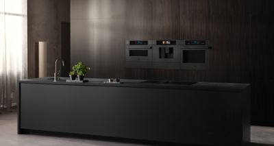 Deze moderne keuken toont een strak zwart kookeiland, een stijlvolle kraan en ingebouwde Siemens ovens tegen een donkere houten wand, terwijl levendige groene planten een verfrissend element toevoegen.