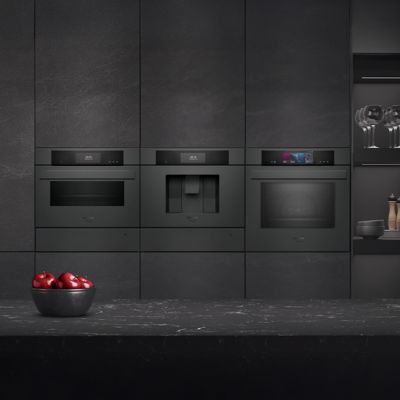 Questa cucina moderna &egrave; dotata di una serie di elettrodomestici, tra i quali un forno e una macchina da caff&egrave;, integrati in una parete scura testurizzata, mentre le mele rosse in una ciotola sul piano di lavoro danno un tocco di colore all'insieme.