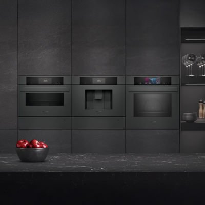 Deze moderne keuken beschikt over zwarte inbouwapparaten, waaronder een oven en koffiezetapparaat, tegen een donkere textuurwand, terwijl een schaal met rode appels een vleugje kleur toevoegt op het aanrecht.