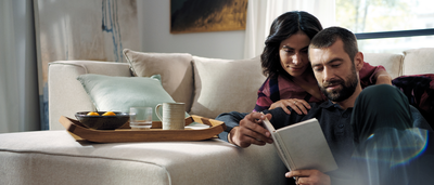 Eine gem&uuml;tliche Wohnzimmerszene zeigt ein Paar auf einem weichen, beigen Sofa, das gemeinsam ein Buch liest. Daneben steht ein Tablett mit Obst und Getr&auml;nken.