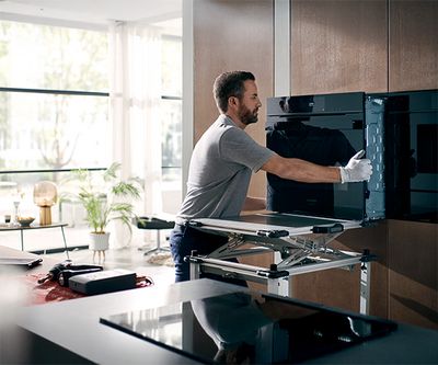 Una persona con guantes ajusta un horno empotrado en una cocina moderna, rodeada de luz y vegetaci&oacute;n.