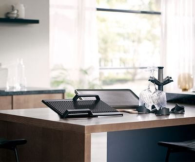 Una encimera de cocina moderna con una sart&eacute;n para grill, una bandeja de horno y copas de vino sobre un escurridor de platos, con la luz del sol filtr&aacute;ndose por las ventanas.