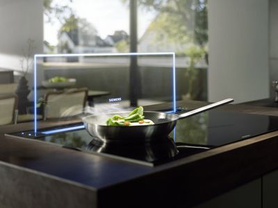 A sleek kitchen with a modern induction cooktop featuring a pan of saut&eacute;ing vegetables, illuminated by natural light from a nearby window.