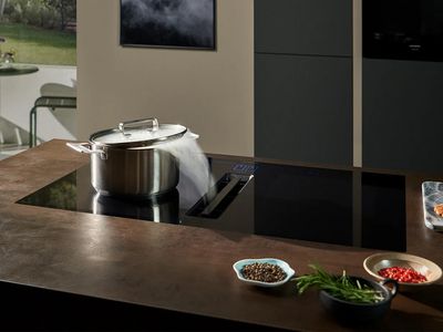 A stainless steel pot with a lid boils on a sleek black cooktop, with steam rising, surrounded by herbs and spices.