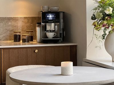 A Siemens coffee machine on a kitchen counter, with a coffee cup being brewed, and a dining table with a candle and floral arrangement in the foreground.