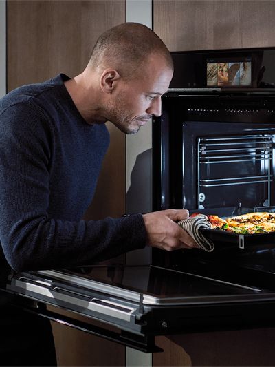 A person pulls a tray of cooked food from a modern oven, wearing a striped oven mitt and standing in a sleek kitchen setting.