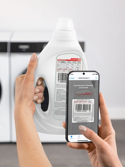 A person holds a laundry detergent bottle while scanning its barcode with a smartphone app in a laundry room setting.
