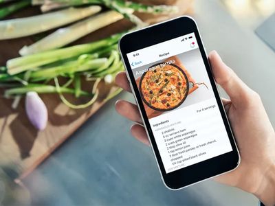 A hand holding a smartphone displaying a recipe for 'Asparagus tortilla' with fresh asparagus and a shallot on a cutting board in the background.