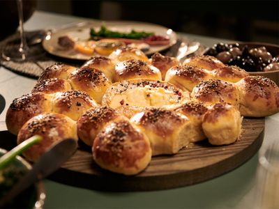 A round wooden board with pull-apart bread rolls surrounding a baked cheese, served on a table with other dishes.