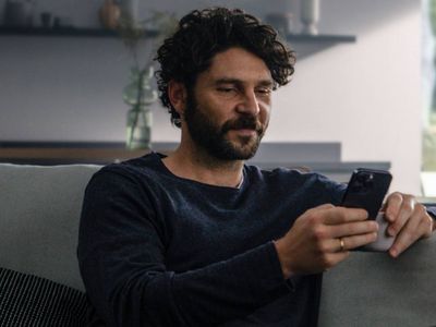A man with curly hair and a beard sitting on a sofa, looking at his smartphone.