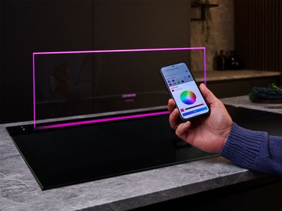 A hand using a smartphone app to control the magenta lighting of a Siemens studioLine downdraft extractor fan and induction hob.