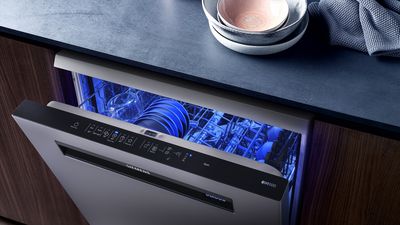 A Siemens dishwasher with its door ajar, revealing a blue-lit interior with dishes, next to a dark countertop with bowls and a cloth.