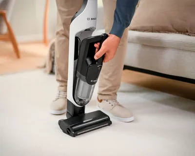 Човек, използващ безжична прахосмукачка Bosch върху плюшен килим в модерна всекидневна.