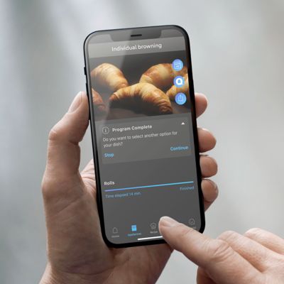 Ruka drž&iacute; chytr&yacute; telefon zobrazuj&iacute;c&iacute; aplikaci pro vařen&iacute;, kter&aacute; ukazuje dokončen&aacute; nastaven&iacute; pečen&iacute; croissantů a v&yacute;zvu k pokračov&aacute;n&iacute; nebo zastaven&iacute;.   