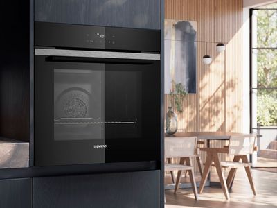 En moderne Siemens sort indbygningsovn monteret i et gr&aring;t k&oslash;kkenskabe ved siden af en lys spisestue.