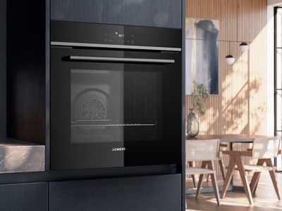 En moderne Siemens sort indbygningsovn monteret i et gr&aring;t k&oslash;kkenskabe ved siden af en lys spisestue.