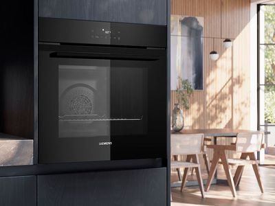 En moderne Siemens sort indbygningsovn monteret i et gr&aring;t k&oslash;kkenskabe ved siden af en lys spisestue.