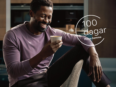 En person i en lavendelf&auml;rgad l&aring;ng&auml;rmad skjorta h&aring;ller en kopp, sittande bekv&auml;mt i ett modernt k&ouml;k.