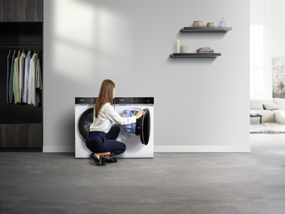 Eine Frau h&auml;lt ein Kleidungsst&uuml;ck neben einer wei&szlig;en Siemens Frontlader‑Waschmaschine und einem passenden Trockner in einem modernen Ankleidezimmer. 