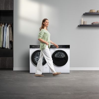 Een vrouw loopt langs een Siemens iQ700-wasmachine en -droger in een lichte wasruimte.
