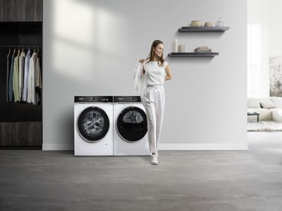 Eine Frau h&auml;lt ein Kleidungsst&uuml;ck neben einer wei&szlig;en Siemens Frontlader‑Waschmaschine und einem passenden Trockner in einem modernen Ankleidezimmer.