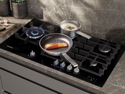 A black Siemens gas hob with blue flames, cooking a salmon fillet in a pan and a pot of creamy soup, set on a dark marble countertop.