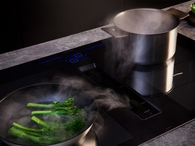 A black Siemens induction cooktop with blue digital controls, featuring an integrated downdraft extractor actively pulling steam from a pot of boiling water and a pan of steaming broccoli.