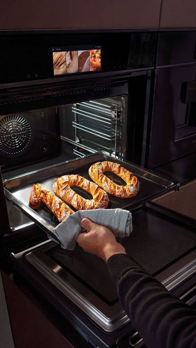 A person removes a tray with freshly baked bread shaped like the number 100 from a modern oven, showcasing a sleek kitchen design