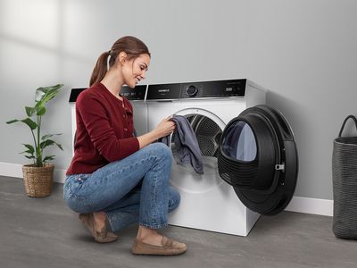 Eine Frau l&auml;dt W&auml;sche in einen wei&szlig;en Siemens Trockner mit schwarzer T&uuml;r f&uuml;r perfekte Trocknungsergebnisse. 