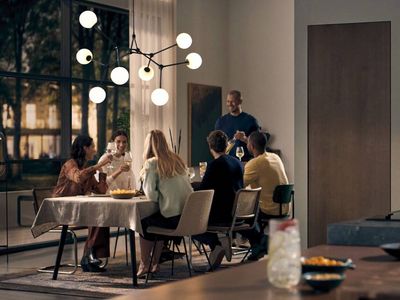 A group of friends enjoys a dinner party in a modern open-plan living space, with a dining table on the left and a sleek kitchen featuring integrated Siemens ovens, a coffee machine, and a microwave on the right.
