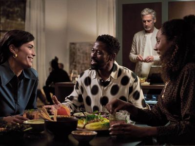 A diverse group of friends enjoys a lively dinner party, with three people in the foreground engaged in conversation around a table laden with food and drinks.