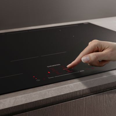           Main manipulant le panneau de commande tactile d'une table de cuisson &agrave; induction noire Siemens sur un plan de travail en pierre.         