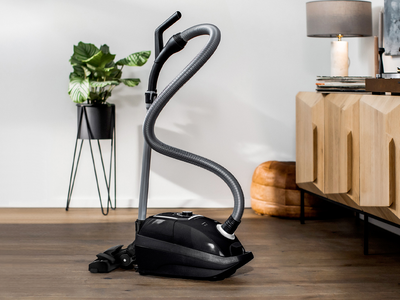 Ein schwarzer Siemens VSZ7X500 Staubsauger mit Beutel steht auf einem Hartboden frei im Raum. Im Hintergrund eine Pflanze in einem Pflanzenst&auml;nder. In das Bild ist ein Testsieger Logo der Stiftung Warentest mit dem Testurteil Gut (2,1) f&uuml;r das Ger&auml;t integriert.