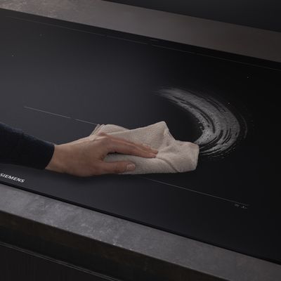           Main essuyant une plaque de cuisson en verre noir avec un chiffon blanc, enlevant un r&eacute;sidu circulaire de savon ou de nettoyant.         