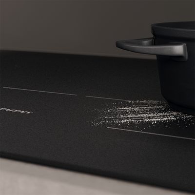 
         Casserole noire sur une plaque &agrave; induction noire avec des granul&eacute;s blancs dispers&eacute;s, probablement du sel ou du sucre, pr&egrave;s du bord de la casserole.
        