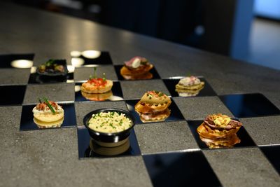 A collection of gourmet appetizers and a bowl of dip arranged on a checkerboard-patterned countertop made of grey stone and reflective black squares.